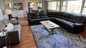 Amplia sala de estar con un sofá seccional de cuero negro, un sillón gris y una mesa de centro. Hay una alfombra estampada en el suelo, y la habitación tiene ventanas grandes que dejan entrar luz natural.
