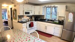 Eine Küche mit weißen Schränken, einer Mikrowelle, einem Herd und einem Kühlschrank. Ein Esstisch mit Blumenmuster-Tischdecke ist ebenfalls vorhanden.