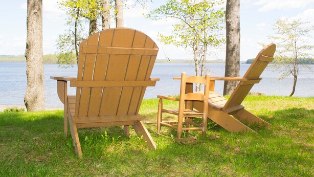 Deux fauteuils Adirondack placés au bord d’un lac, entourés d’arbres, et offrant une vue dégagée sur l’eau.