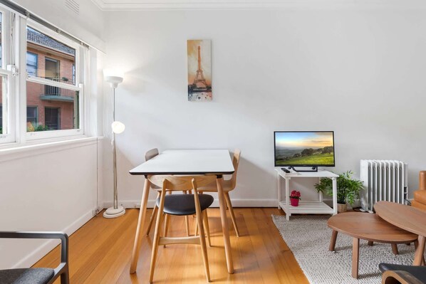 Un salon moderne comprenant une table à manger, des chaises, un téléviseur, un radiateur et une fenêtre donnant sur un immeuble.