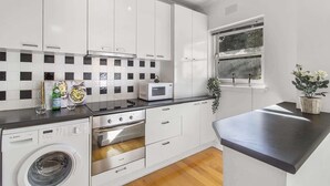 A modern kitchen with white cabinets, a microwave, a washing machine, and a window with a view. - Comfy One-Bedroom Unit Between City and Airport (Travancore)
