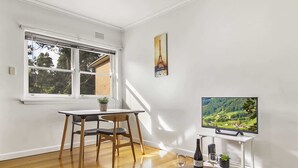 A room with a dining table, chairs, a TV on a stand, a bottle of wine, and glasses. - Comfy One-Bedroom Unit Between City and Airport (Travancore)