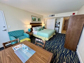 A hotel room with a bed, a sofa, a table with water bottles, a microwave, and a TV. - Oceanfront 2 Room Suite with Resort Amenities. (Hollywood)