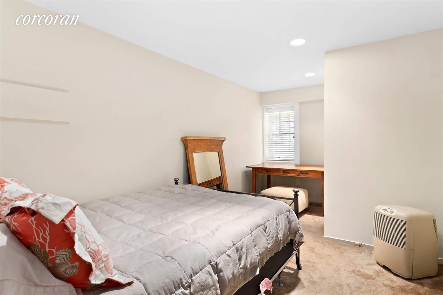 A bedroom with a bed, a wooden desk, a chair, a mirror, and a standing heater.