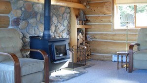 Interior de una cabaña de troncos con pared de piedra, chimenea, sofá y sillón.