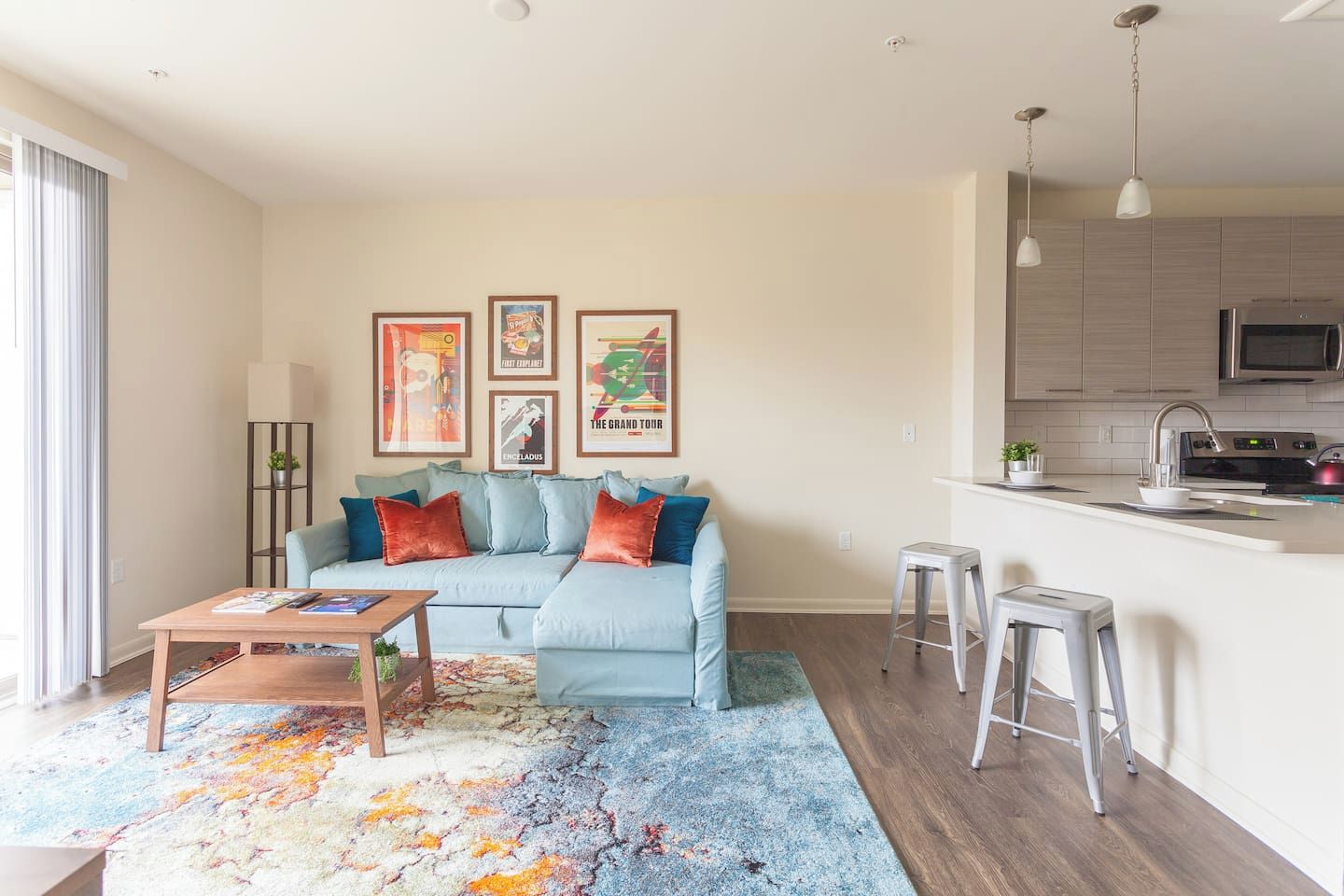 A living room with a sofa, coffee table, and colorful rug. There is a kitchen area with a microwave and a sink.