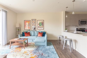Sala de estar con sofá, mesa de centro y alfombra colorida. Hay una zona de cocina con microondas y fregadero.