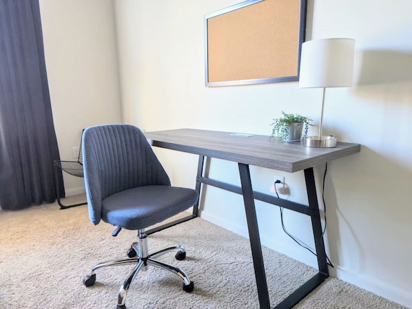 Una habitación con escritorio, silla, lámpara y un tablón de corcho en la pared.