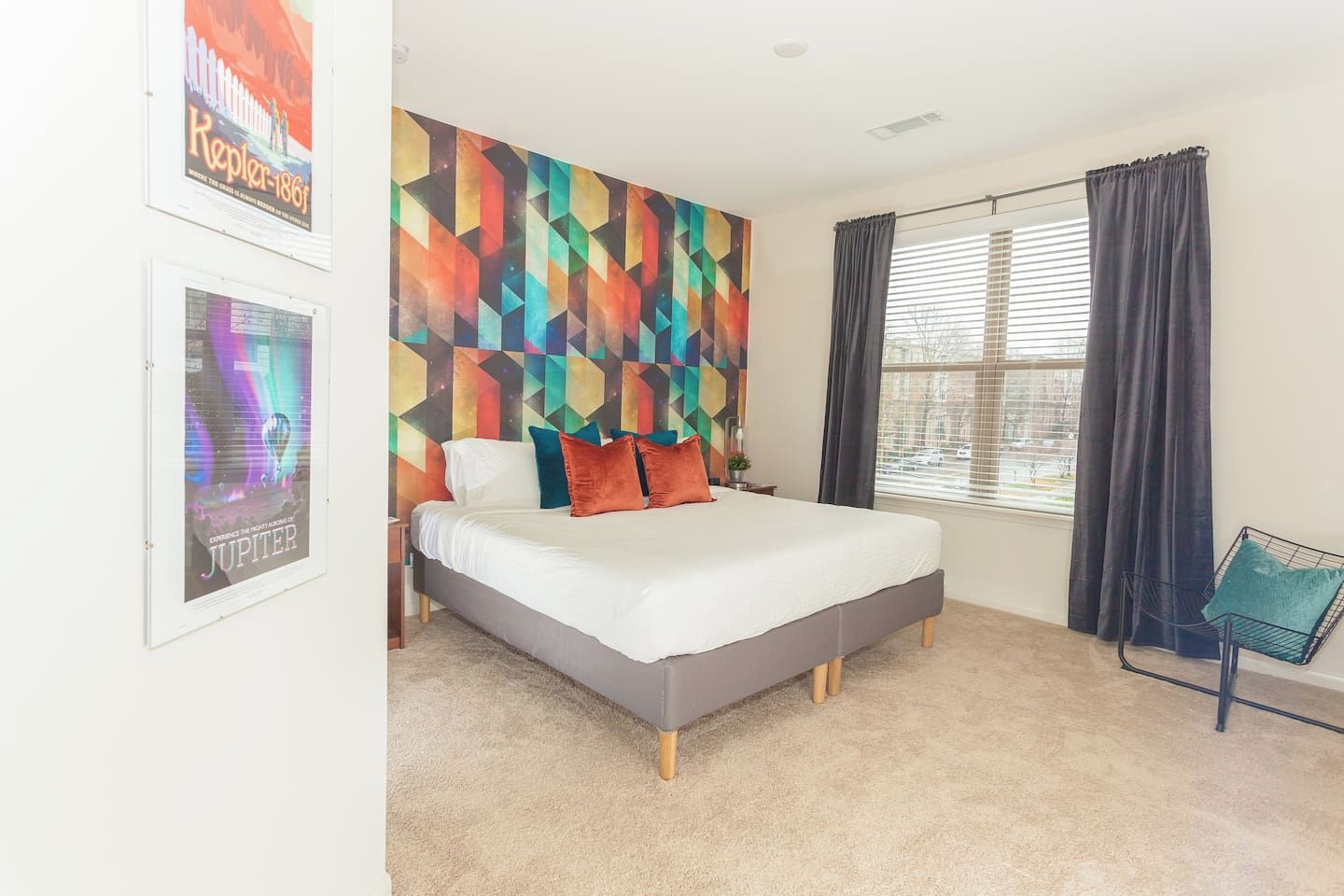 A bedroom with a bed, a colorful geometric wall art, a window with curtains, and a framed poster on the wall.