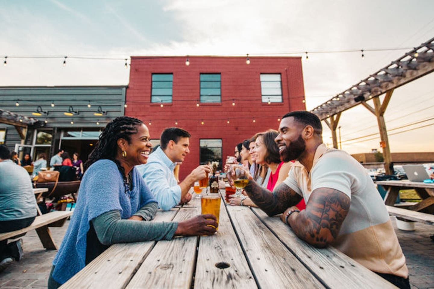 A group of people toasting at a wooden table outdoors with a red building in the background.