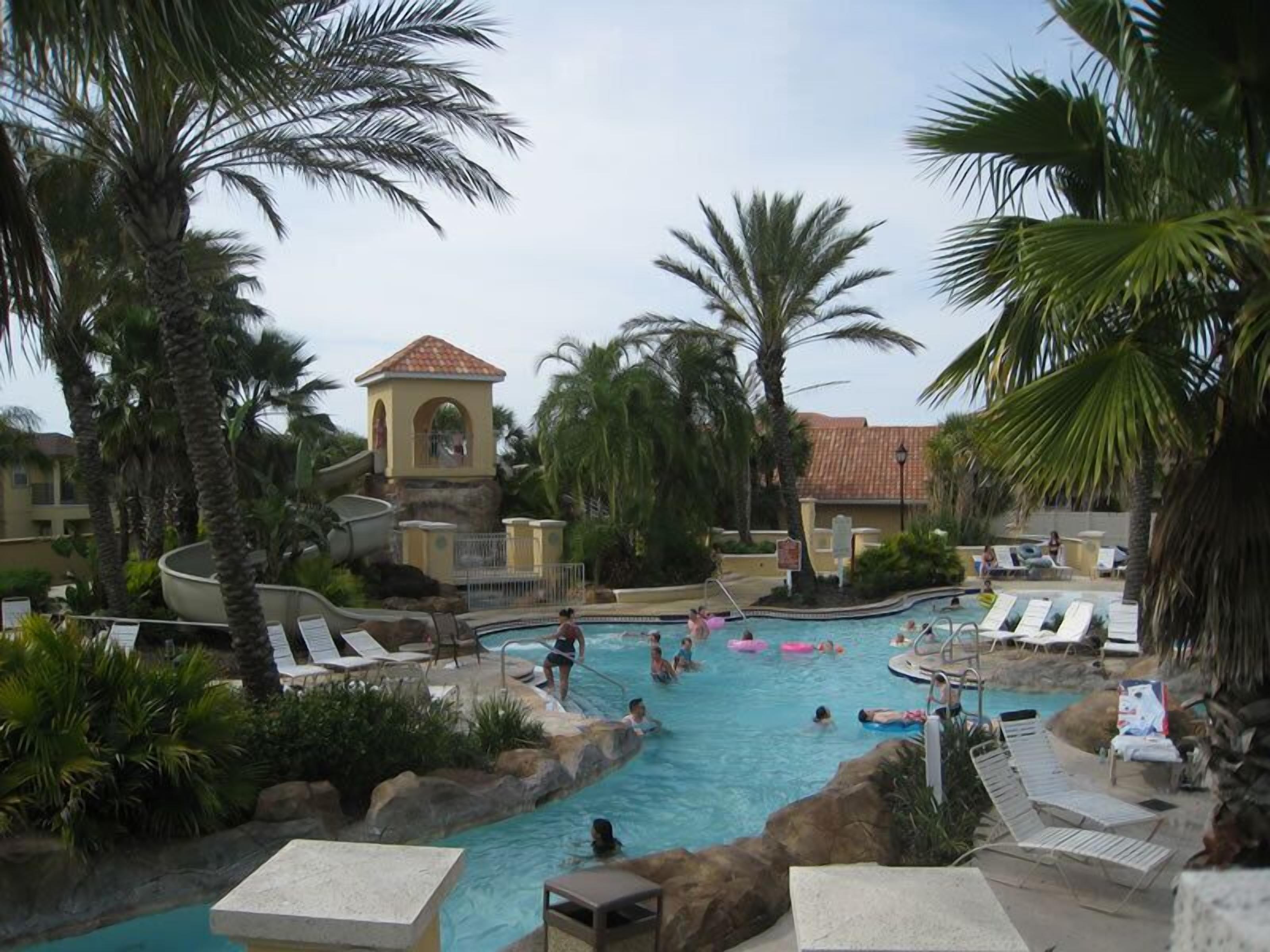 Ein Poolbereich eines Resorts mit Palmen, Liegestühlen und einem kleinen Gebäude mit einem Ziegeldach.