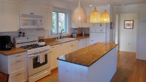 A modern kitchen with white cabinetry, a central island, a stove, a microwave, and a refrigerator. There are pendant lights above the island. - Attractive 3BR Cape Home Close to Indian Neck Beach (1430) (Wellfleet)