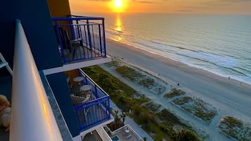 A resort balcony with a view of the ocean at sunset, a pool, and lounge chairs.