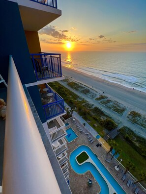Un balcon d’hôtel avec vue sur l’océan au coucher du soleil, une piscine et des chaises longues.