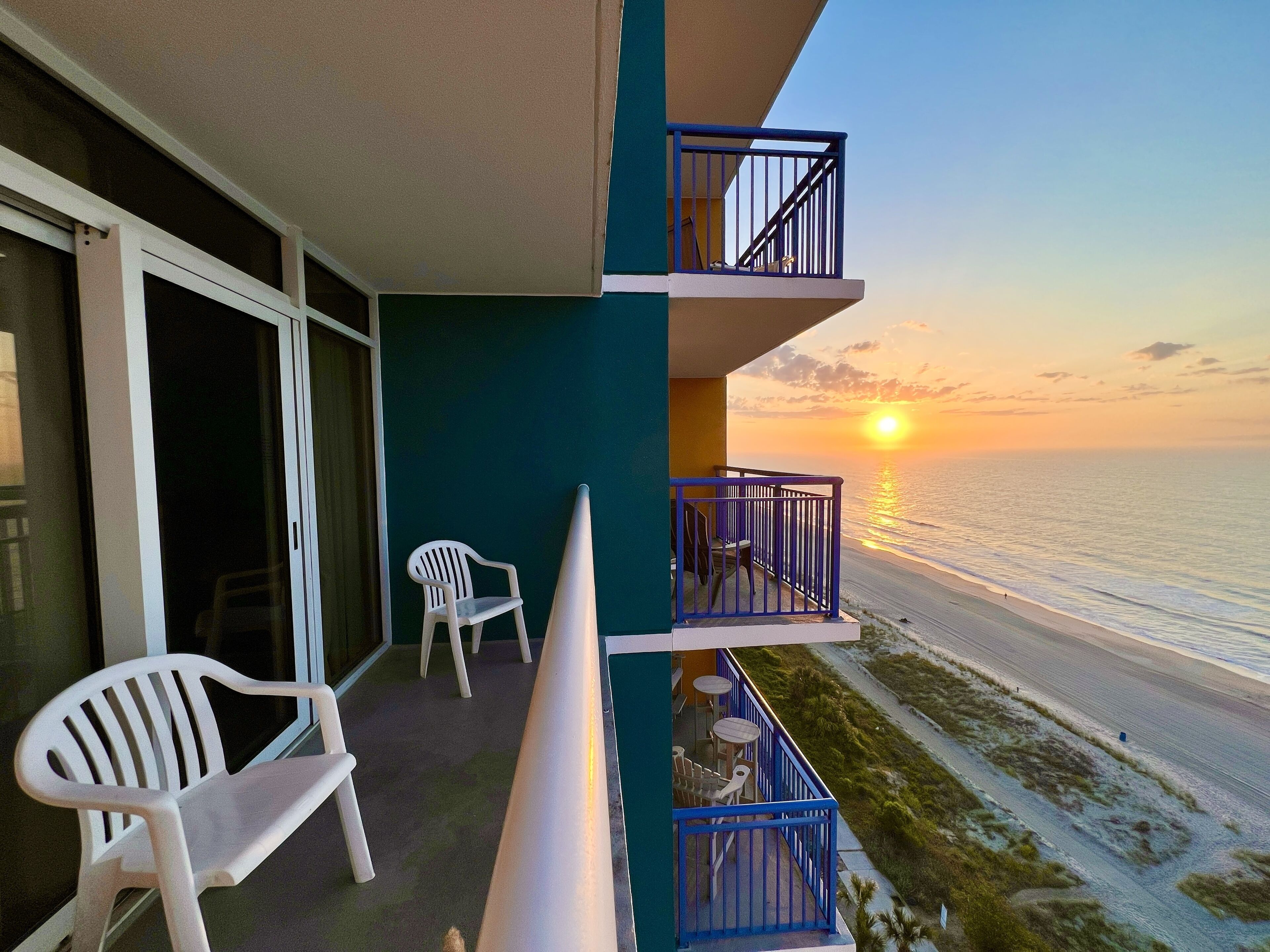 A balcony with a view of the sunset over the ocean, a white chair, and a table with chairs.