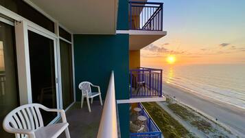 A balcony with a view of the sunset over the ocean, a white chair, and a table with chairs.