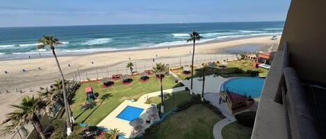 Une vue sur la plage depuis un balcon donnant sur une piscine, des palmiers et une plage de sable.