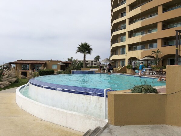 Una piscina rodeada por un edificio con balcones, palmeras y una pequeña construcción con techo de tejas.