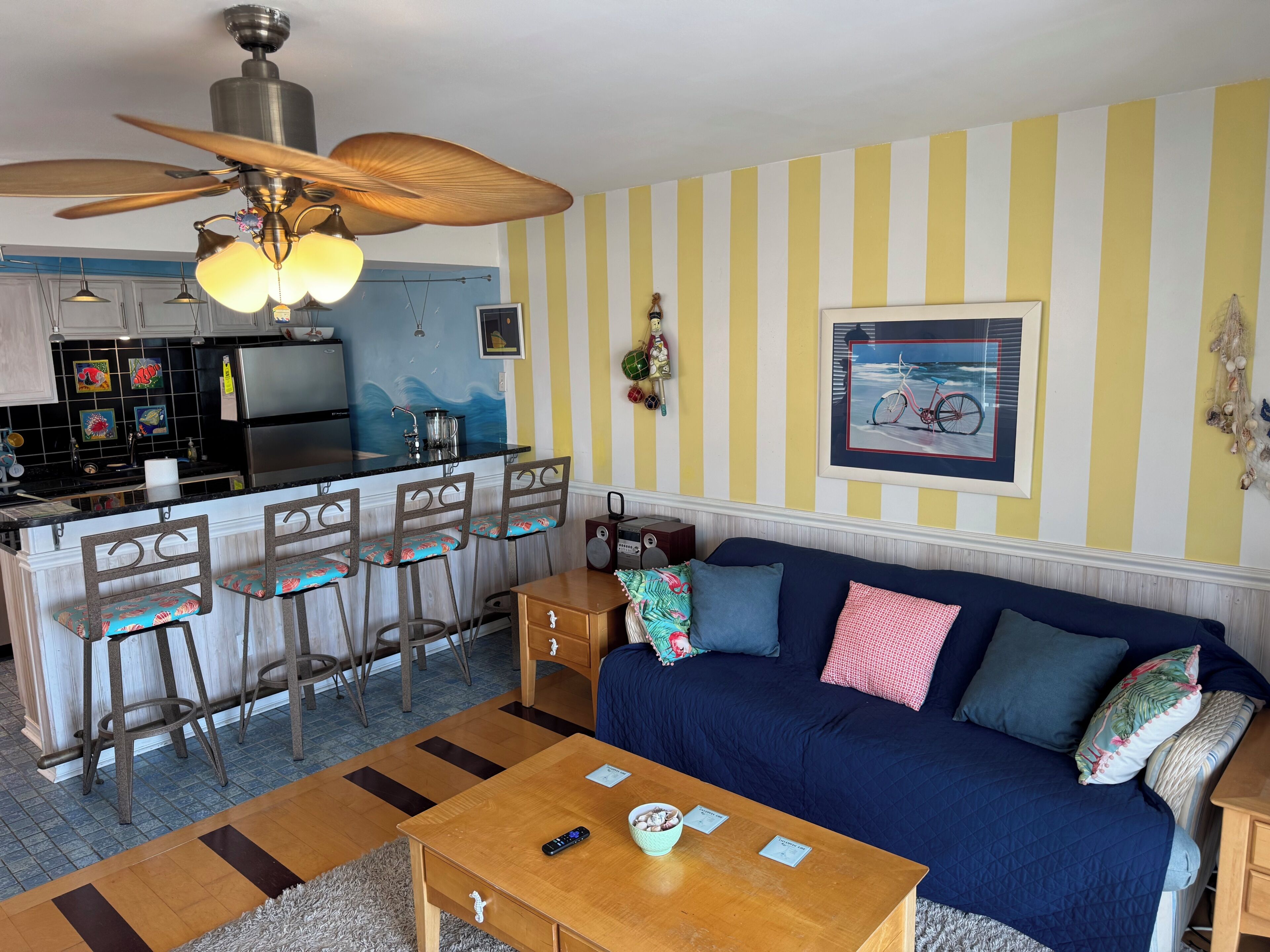 A kitchen with a central island, bar stools, a ceiling fan, and a dining area with a sofa and coffee table.