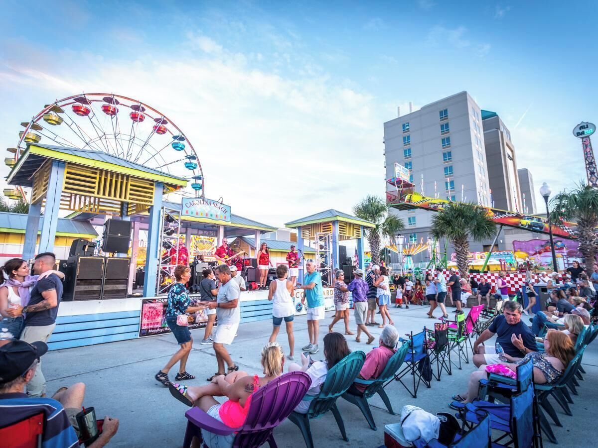 A bustling outdoor event with a Ferris wheel, a stage with performers, and a crowd of people.