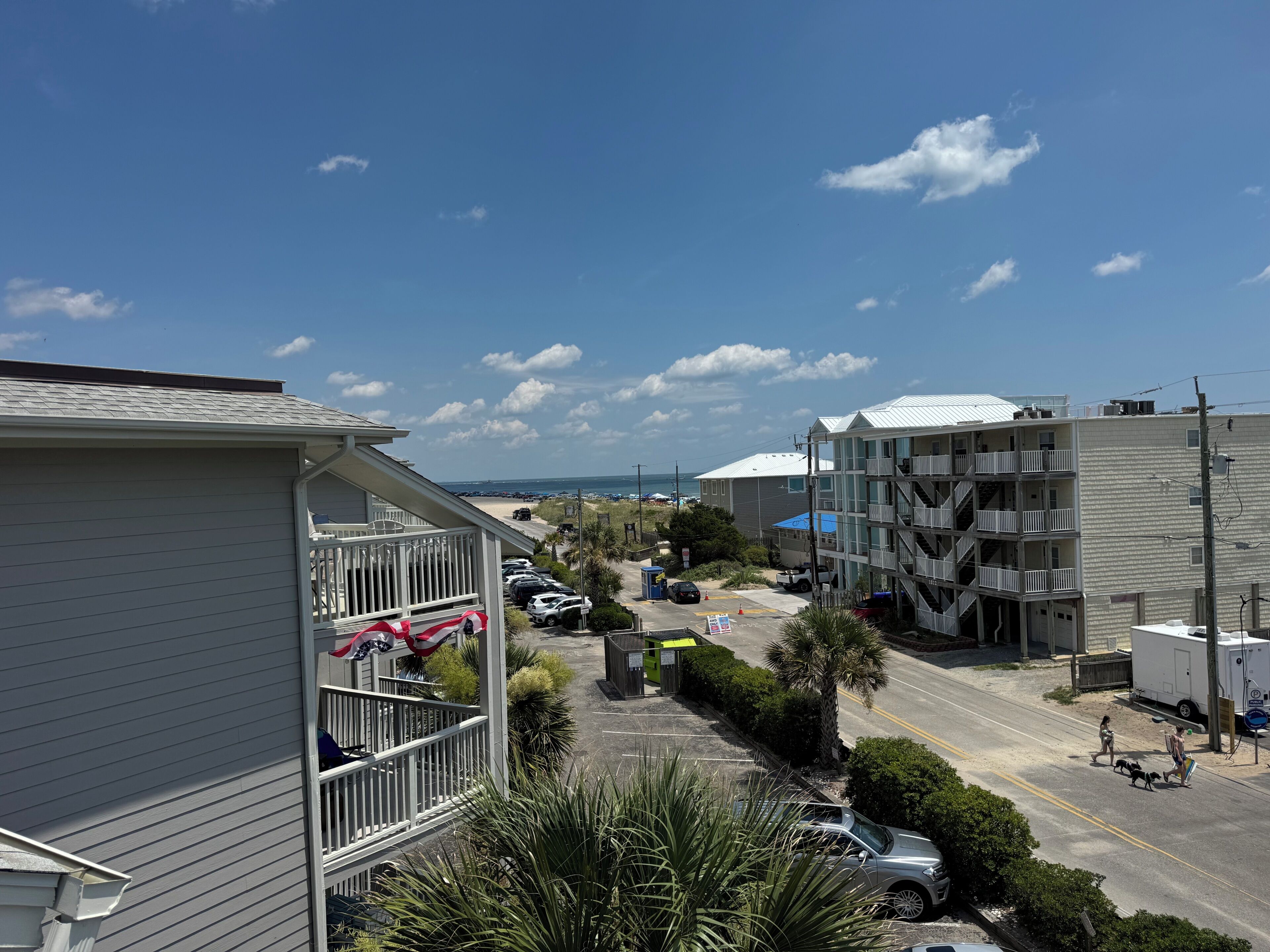 A residential area with a parking lot, a beach in the background, and a multi-story building with balconies.