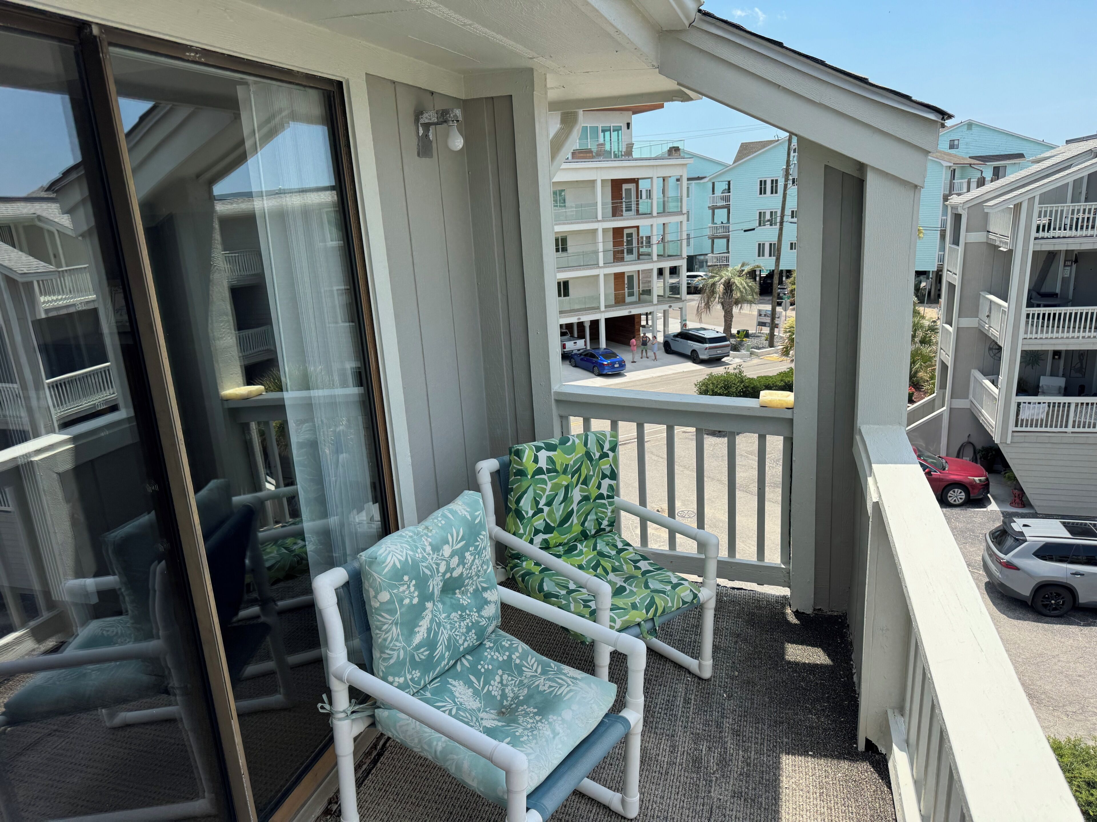 A balcony with two chairs, a glass door, and a view of adjacent buildings and parked cars.