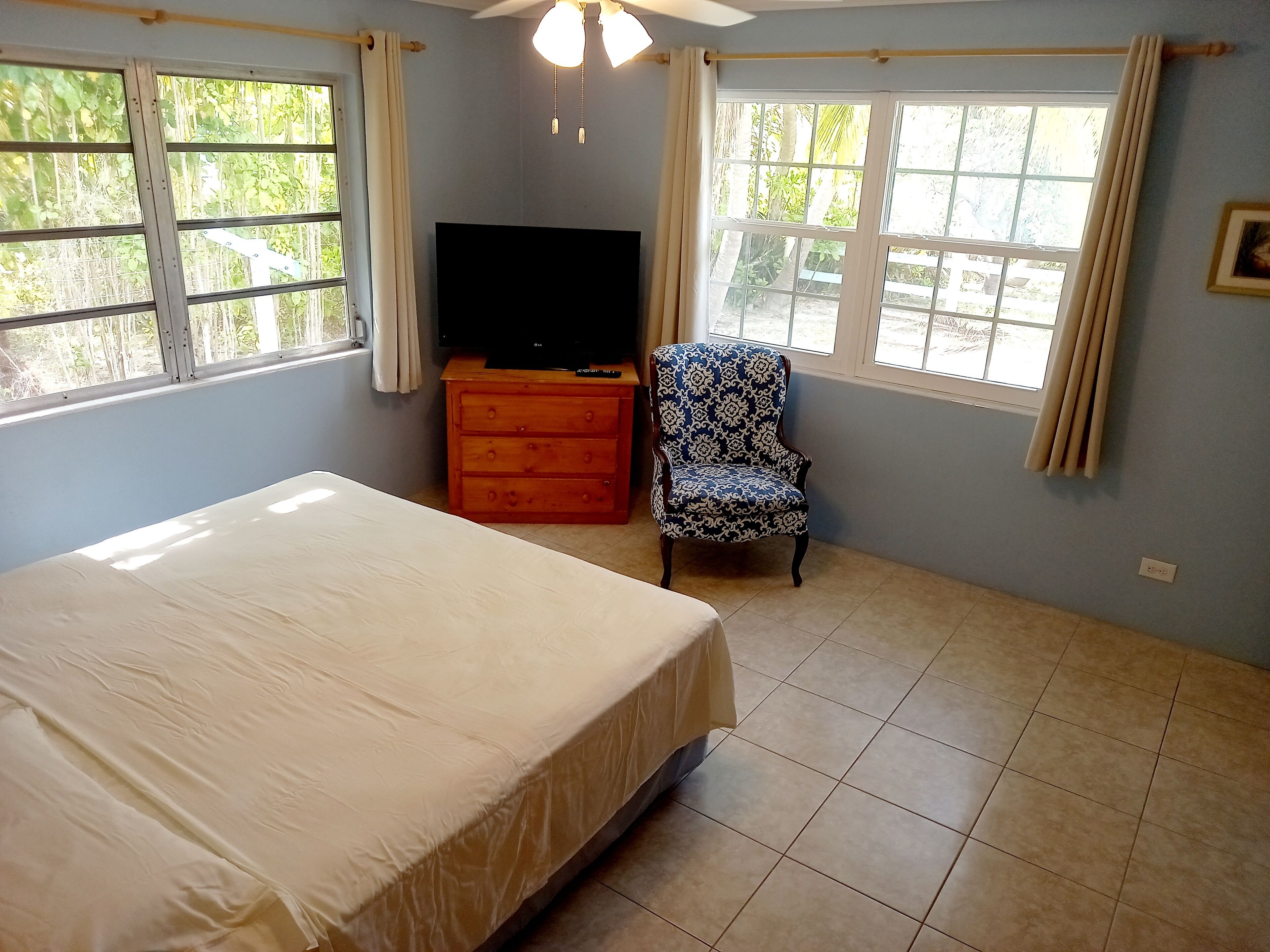 A room with a bed, a wooden dresser, a patterned armchair, a television, and large windows with curtains.