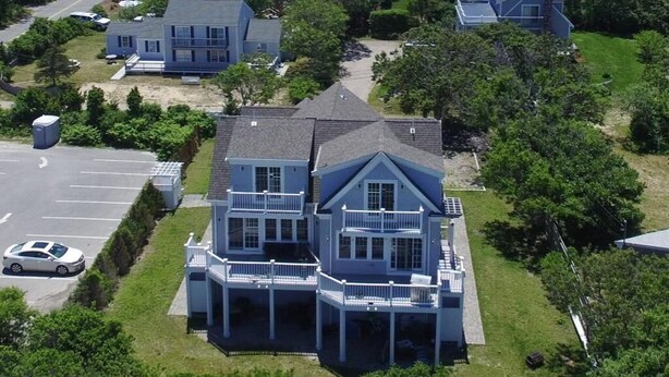 A large, two-story house with a balcony and a covered porch, surrounded by greenery and a parking lot. - Accommodates 12 Guests Overlooking Cape Cod Bay, 6 BR/6 BTH (2048) (Dennis)