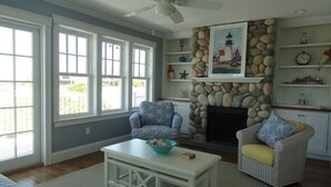 A living room with a stone fireplace, two armchairs, a coffee table, and a ceiling fan. - Accommodates 12 Guests Overlooking Cape Cod Bay, 6 BR/6 BTH (2048) (Dennis)