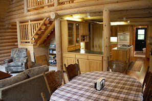 Interior d'una cabana de fusta amb zona de menjador, cuina i escala que condueix a un altell.