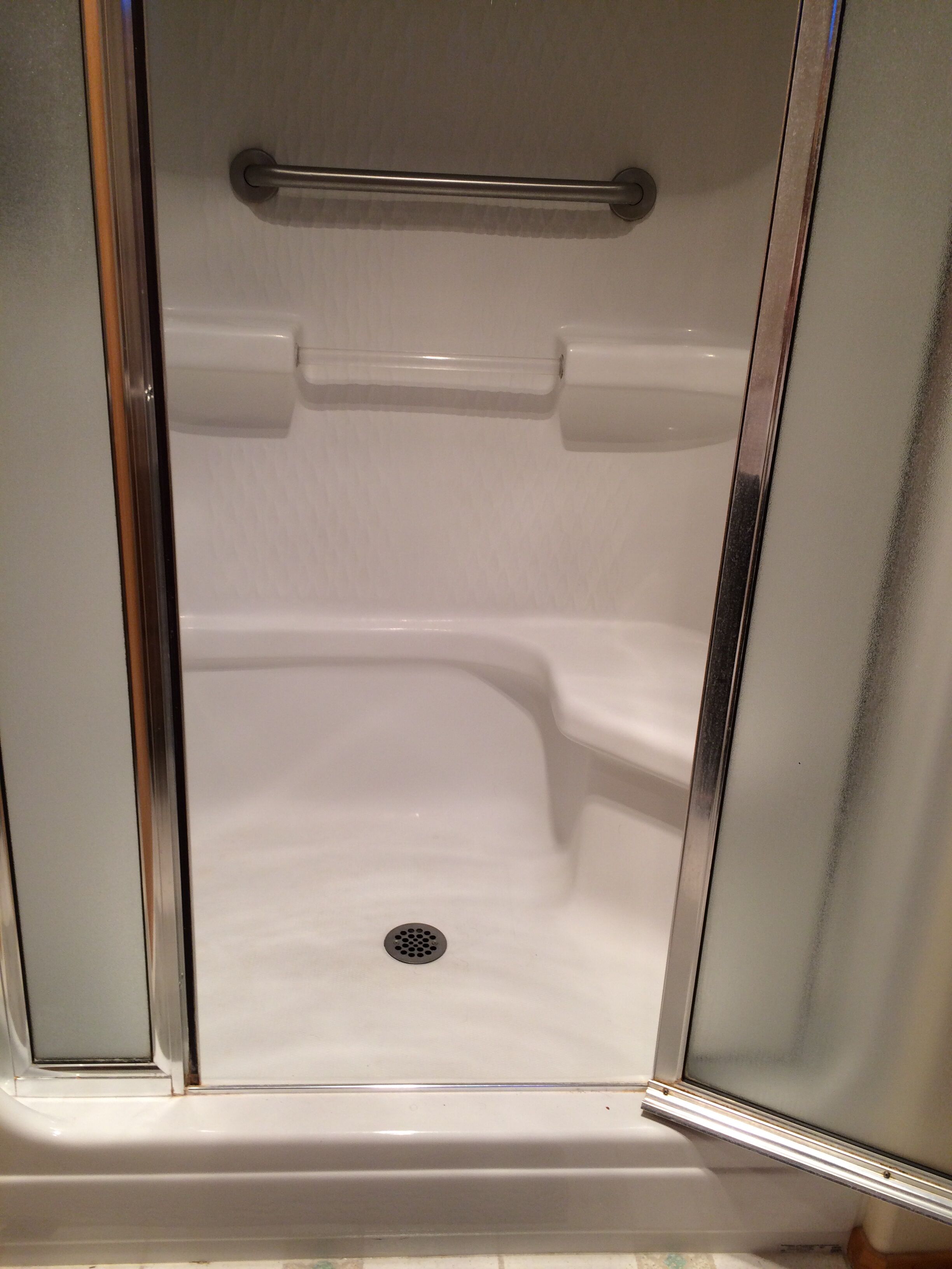 A white, empty shower stall with a metal grab bar and a drain.