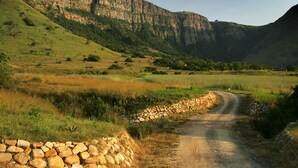 A rural dirt road flanked by stone walls and green fields, leading towards a mountainous backdrop.