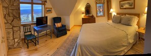 A cozy bedroom with a large bed, a desk with a chair, a dresser, and a window with a view of houses.