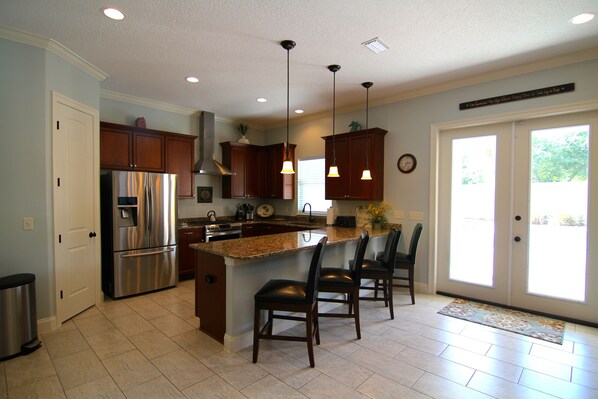 A kitchen with a stainless steel refrigerator, wooden cabinets, a granite countertop, and a bar area with two chairs.