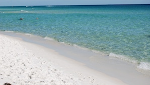 Une plage à l’eau turquoise et limpide, au sable blanc, où quelques personnes se baignent.