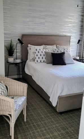A bedroom with a bed, a chair, a small table, and a potted plant.
