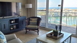 Un salon doté d’un grand téléviseur, d’un meuble bleu, d’un fauteuil à motifs et offrant une vue sur la mer depuis un balcon.