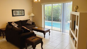 A living room with a dark brown sofa, two wooden side tables, a ceiling fan, and a view of a swimming pool through large glass doors. - Quiet and beautiful place (Kissimmee)