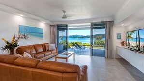 Ein modernes Wohnzimmer mit Ledersofa, Couchtisch und großem Fernseher. Eine Glastür öffnet sich zu einem Balkon mit Blick auf Meer und Berge.