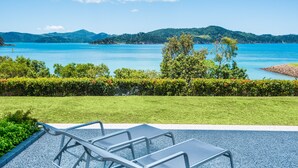 Blick auf eine Küstenlandschaft mit Rasenfläche, einer Sitzgelegenheit mit Metallstühlen und einem klaren, blauen Meer mit weit entfernten Hügeln.