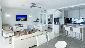 A modern living room with a flat-screen TV, a ceiling fan, and a dining area with a kitchen in the background.