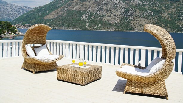 Deux fauteuils de relaxation en osier, dotés de coussins, sont placés sur une terrasse offrant une vue sur un plan d’eau et des montagnes.