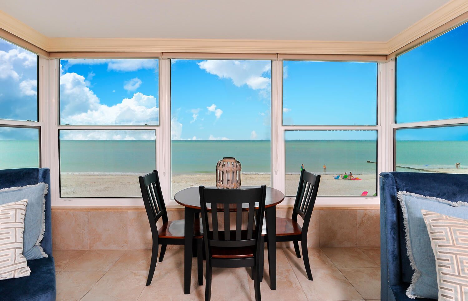 A room with a large window overlooking a beach, a round table with chairs, and blue upholstered seating.