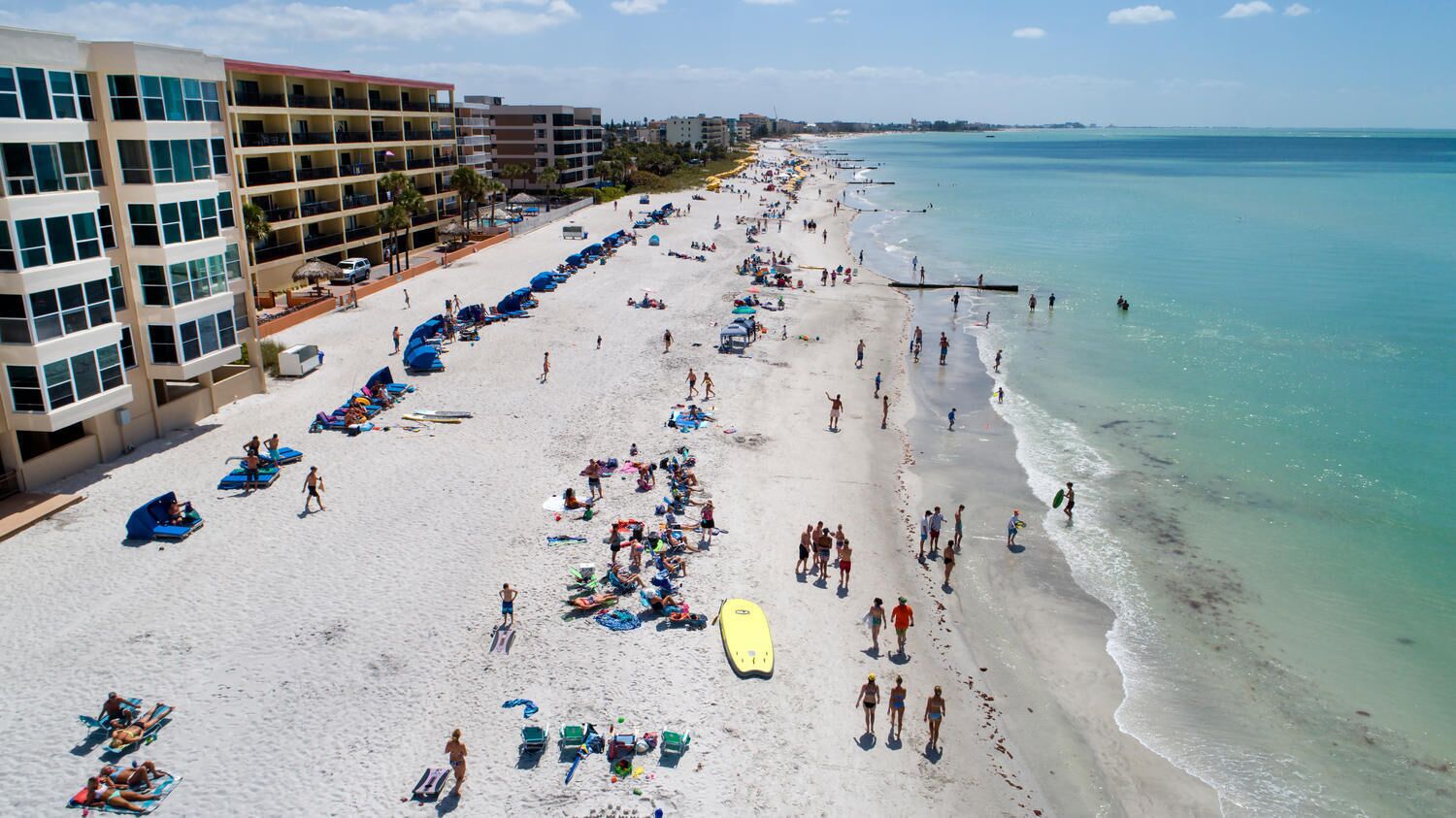 Ein Strand mit mehreren blauen Sonnenschirmen und Liegestühlen, einem gelben Surfbrett sowie einem Gebäude mit mehreren Etagen und Balkonen.