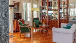 Amplio salón con piso de madera, sillas tapizadas en verde, un sofá blanco y un aparador de vidrio lleno de objetos decorativos.