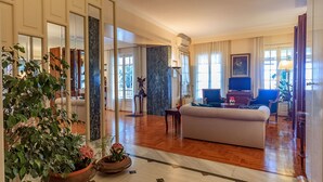 Amplia sala de estar con sofá, televisión y comedor con sillas y mesa. Hay plantas en macetas y un pilar de mármol en la habitación.