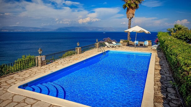 Piscina con vista al mar, rodeada de sillas de descanso y una palmera.