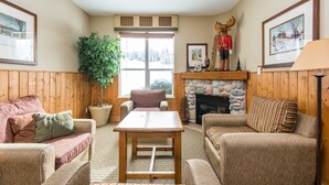 A cozy living room with wooden paneling, a fireplace, a large window, and a mounted moose head. - Large balcony, walk out condo on trails - Pet friendly! (Silver Star Mountain)