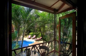 Vista desde un espacio cubierto a una piscina rodeada de palmeras, con sillones de descanso y una mesa con jarra y vaso.
