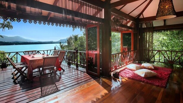 A wooden deck with a table and chairs, a red rug, and a view of a lake and mountains.